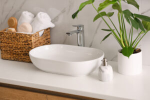 Picture of a bathroom sink, a soap dispenser, a potted plant, and a basket full of towels and other bathroom items.